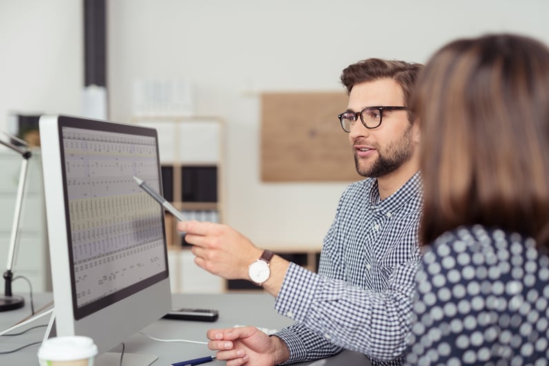 Proficient young male employee with eyeglasses and checkered shirt, explaining a business analysis displayed on the monitor of a desktop PC to his female colleague, in the interior of a modern office-1